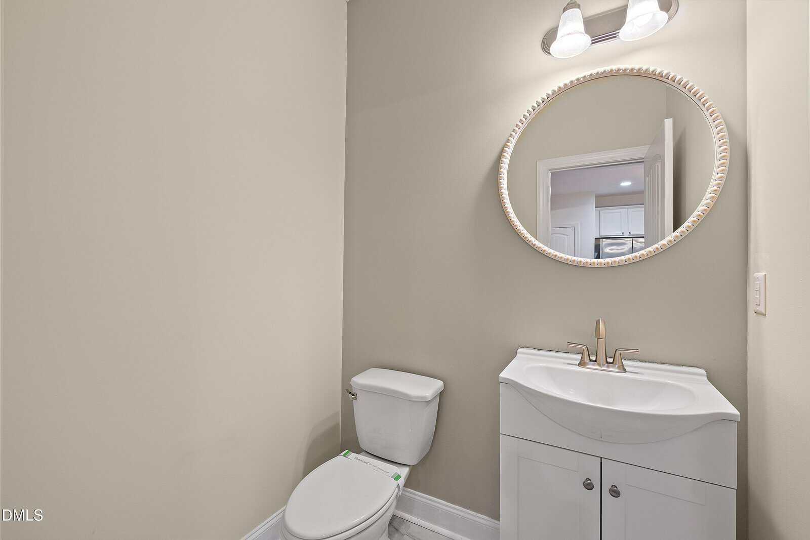 A small bathroom with a white toilet, a white vanity with a curved sink, a gold faucet, an oval framed mirror, and a wall-mounted light fixture above the mirror. The walls are painted beige.