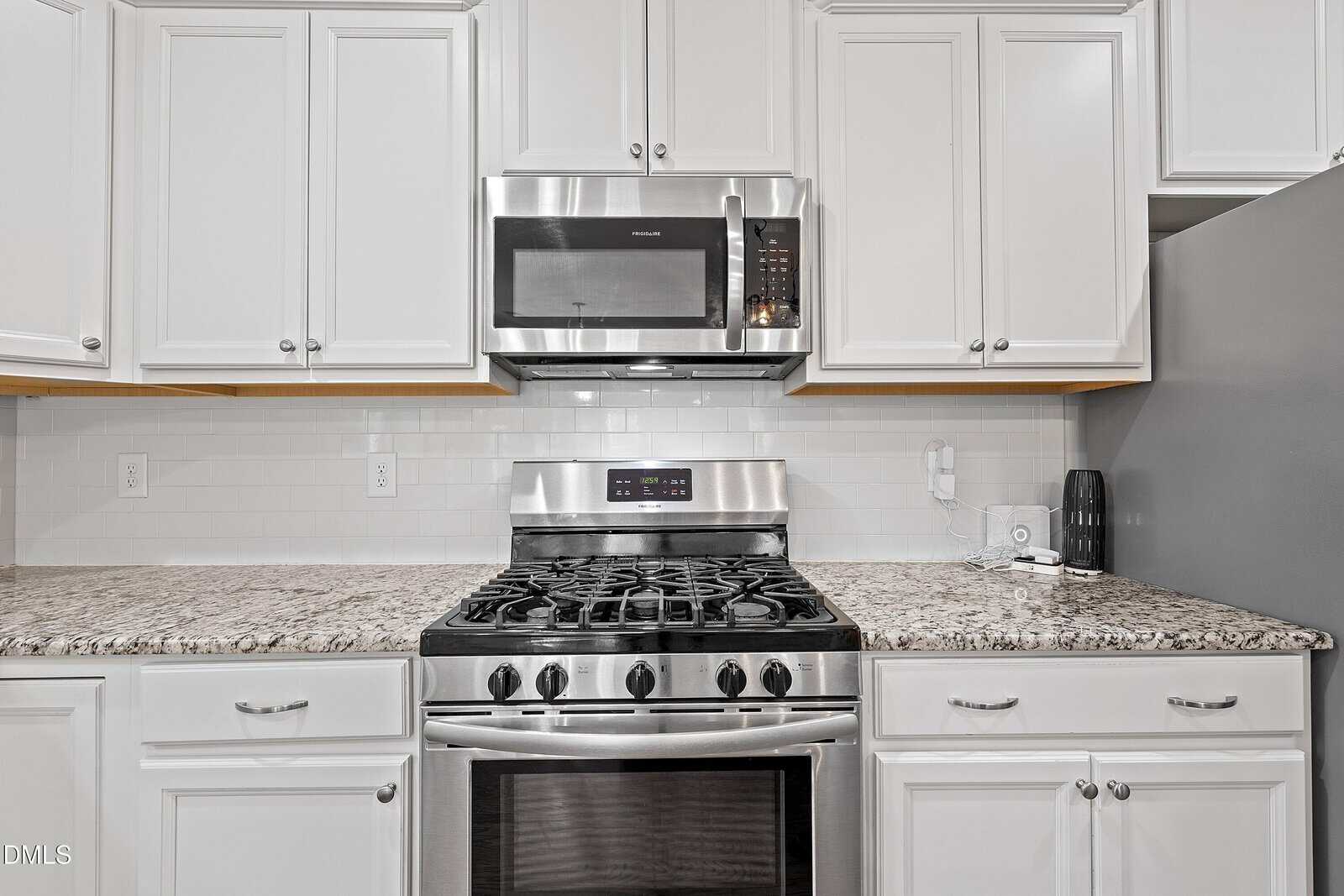 Modern kitchen with white cabinets, granite countertops, a stainless steel gas stove, and a microwave oven above. The backsplash is white tile, and there are small appliances on the right counter.