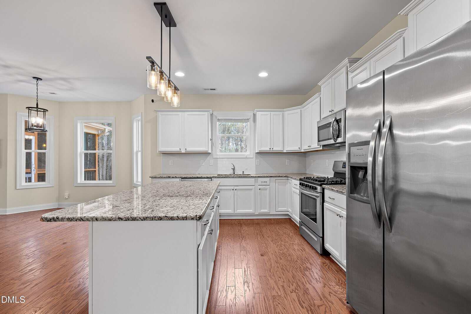 Modern kitchen with white cabinets, granite countertops, stainless steel appliances, wood floors, and island with pendant lights. Dining area with large windows is visible in the background.