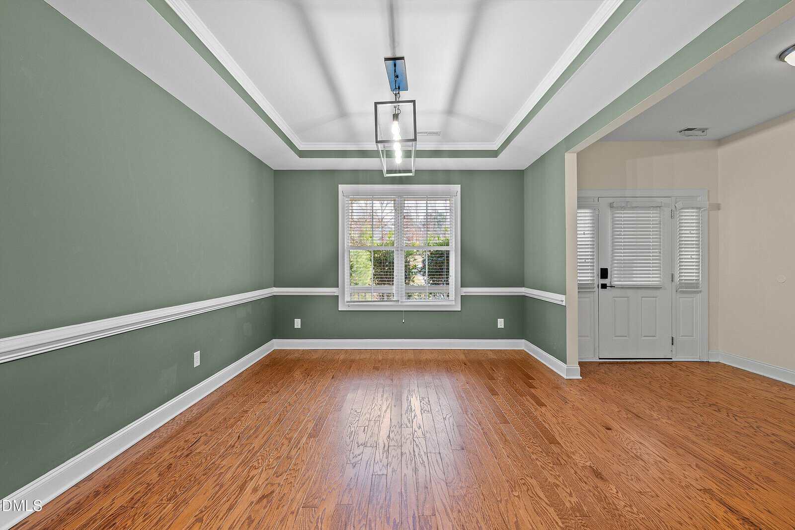 An empty room with green walls, white trim, wood flooring, a large window with blinds, a modern ceiling light fixture, and an adjacent entryway with a white door and sidelights.
