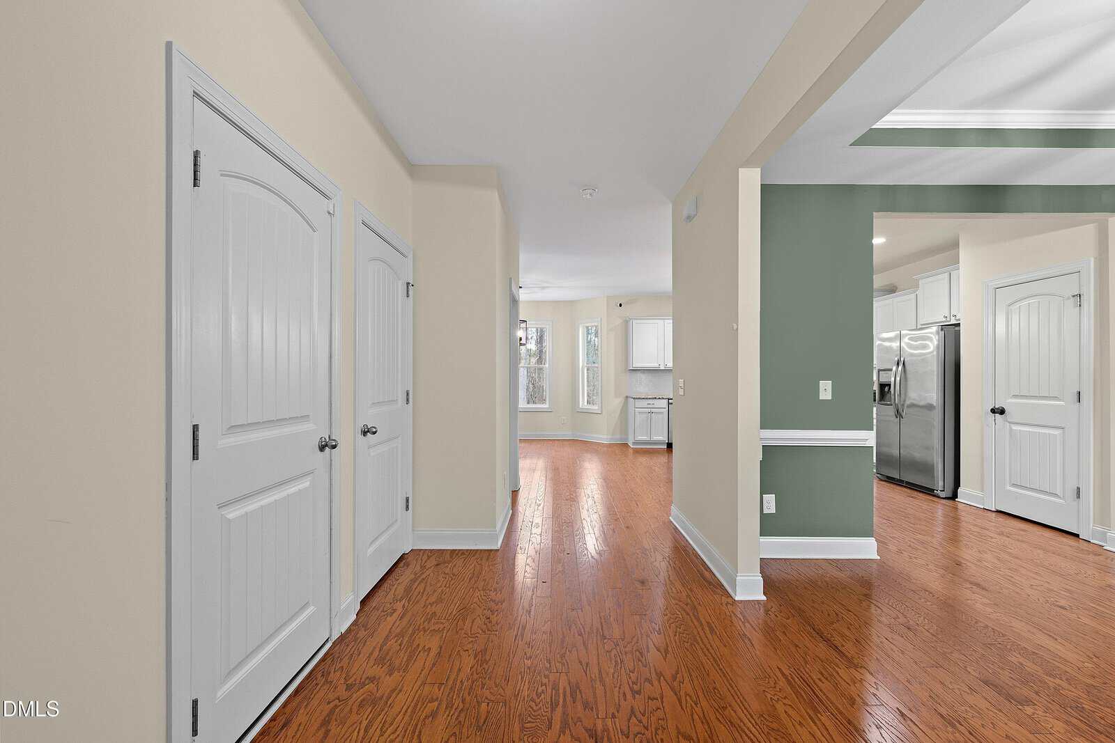 Interior view of a house with light beige walls, hardwood floors, two white paneled doors on the left, and an open kitchen area with stainless steel appliances and green accent walls on the right.