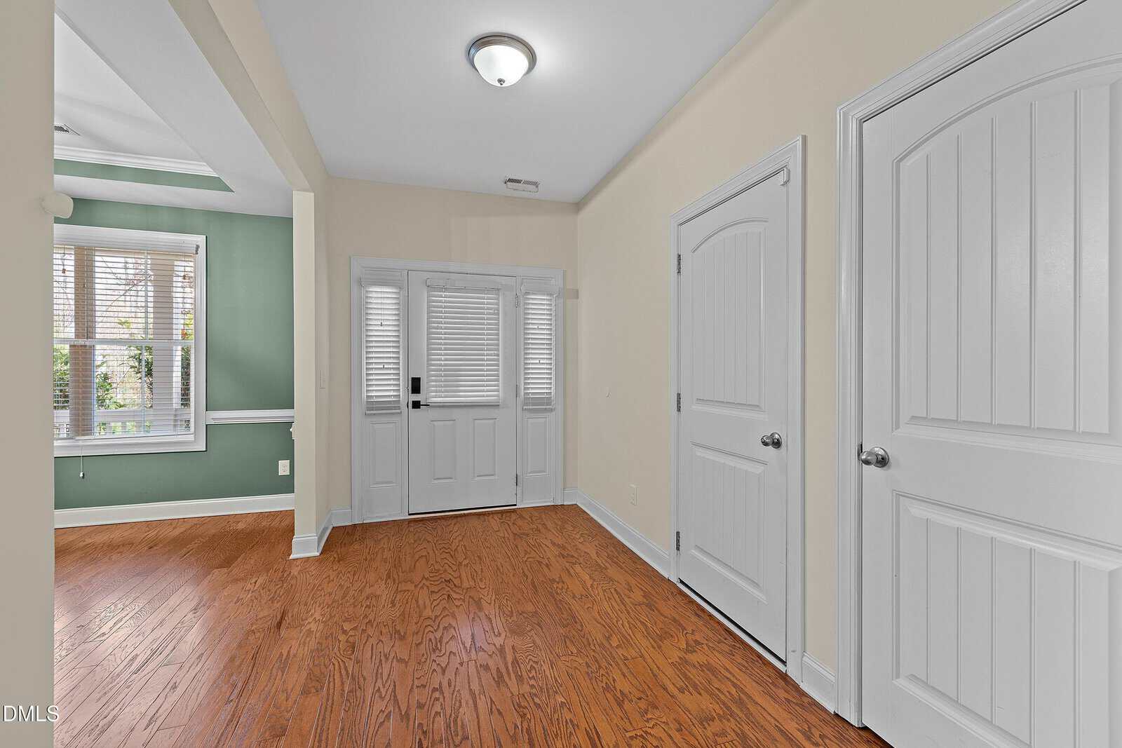 A foyer with wood flooring, beige and green walls, white trim, and two closed white doors. The entry has double front doors with glass panels and side windows, and a chandelier-style ceiling light.