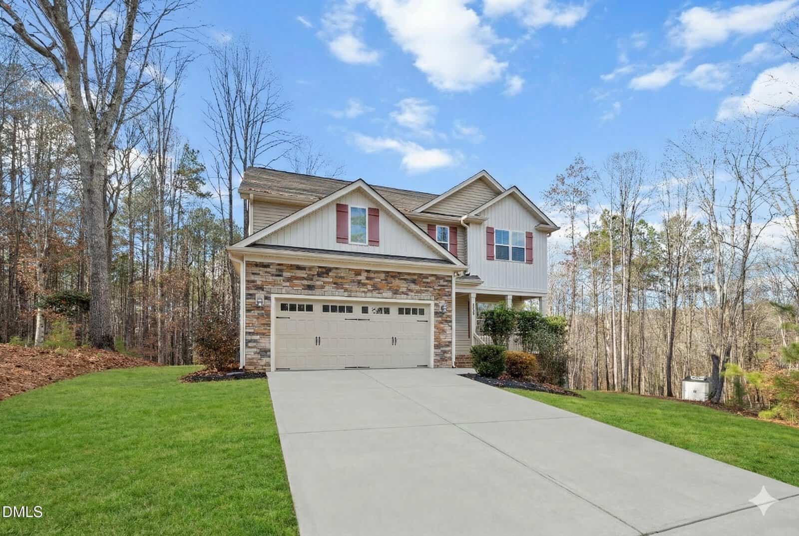 Two-story suburban house with stone and siding exterior, attached two-car garage, concrete driveway, and well-kept lawn. Leafless trees surround the property under a partly cloudy sky.