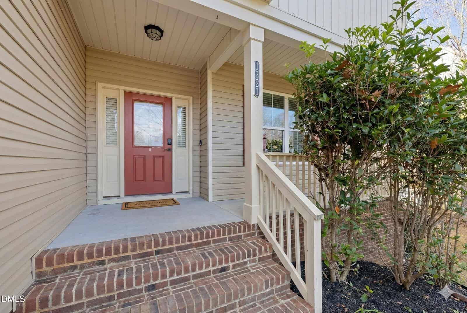A front porch with brick steps, beige siding, and a red door. White railings and columns frame the entry. A welcome mat is at the door, and a green shrub is on the right side of the steps.