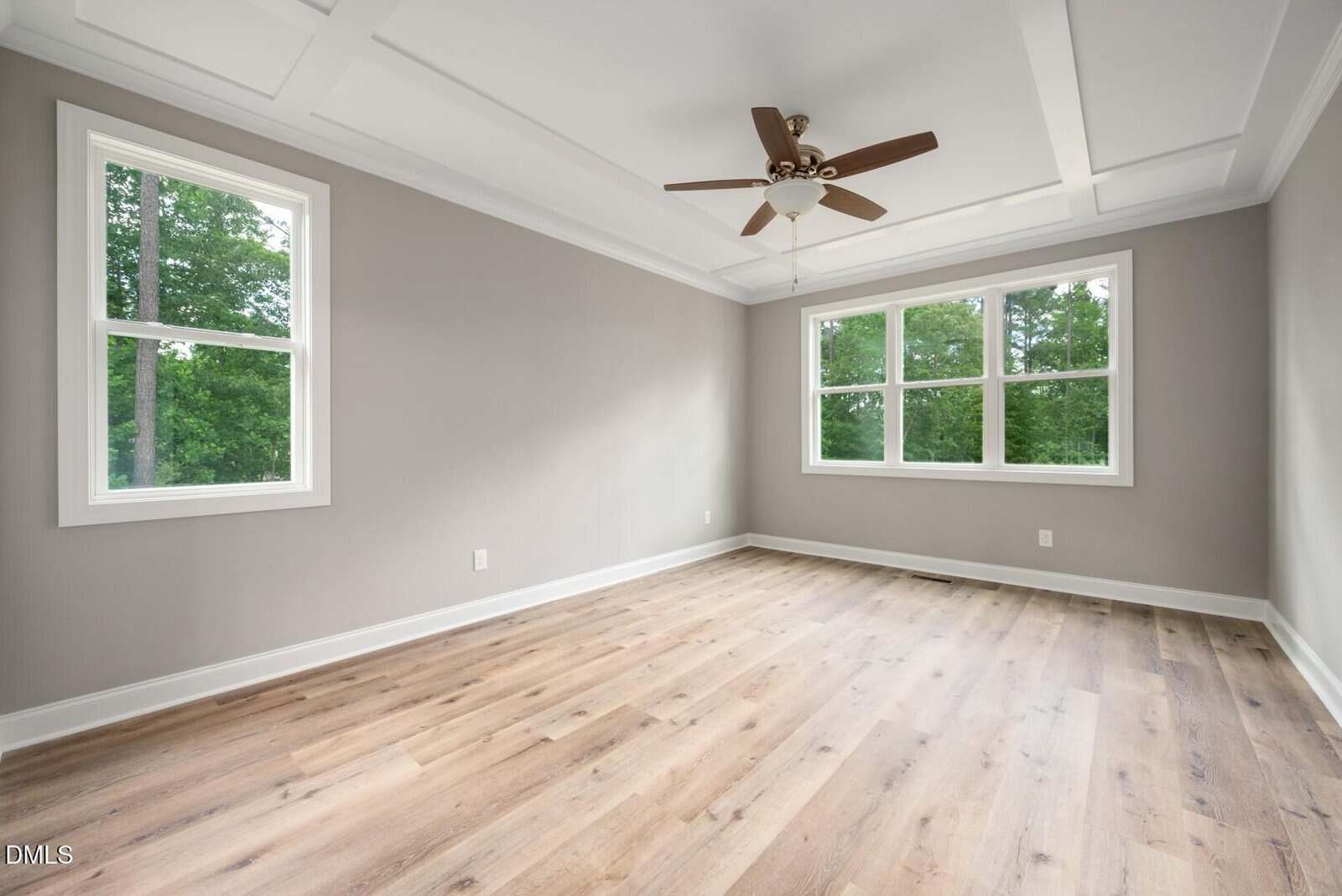 Empty room with light wood flooring, light gray walls, two windows, a ceiling fan with lights, and a tray ceiling with white trim. Trees are visible outside through the windows.