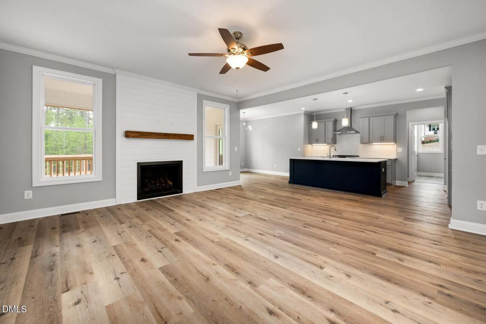 Spacious open-concept living area with light wood floors, gray walls, a ceiling fan, a fireplace with a wooden mantle, and a modern kitchen with an island and pendant lights in the background.