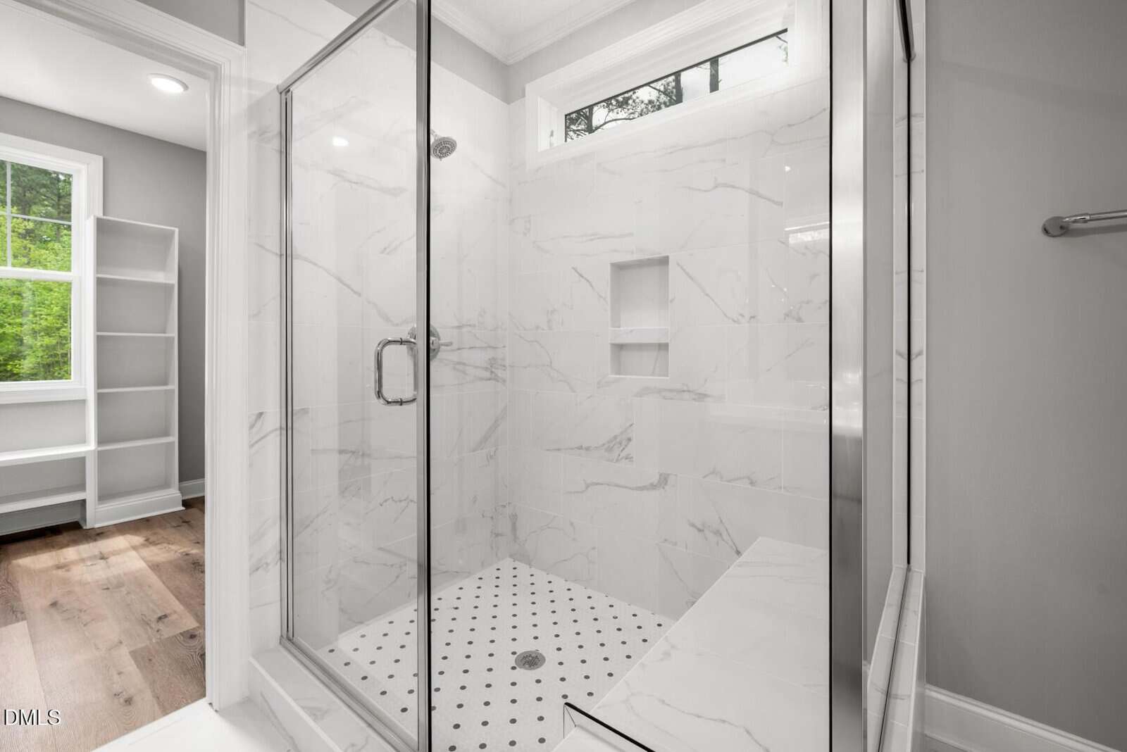 Modern shower with glass doors, marble tile walls, built-in shelves, and a bench. The bathroom has a window with greenery outside and light wood flooring visible in the adjacent room.