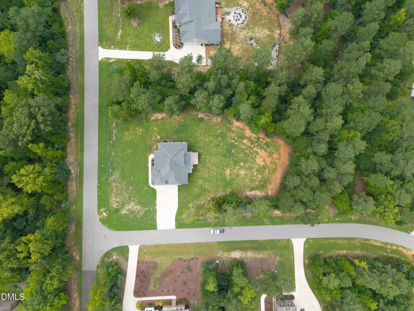 Aerial view of a suburban neighborhood with single-family homes, driveways, and surrounding trees. One house is centered on a grassy lot, with roads bordering two sides. A white vehicle is parked on the lower street.