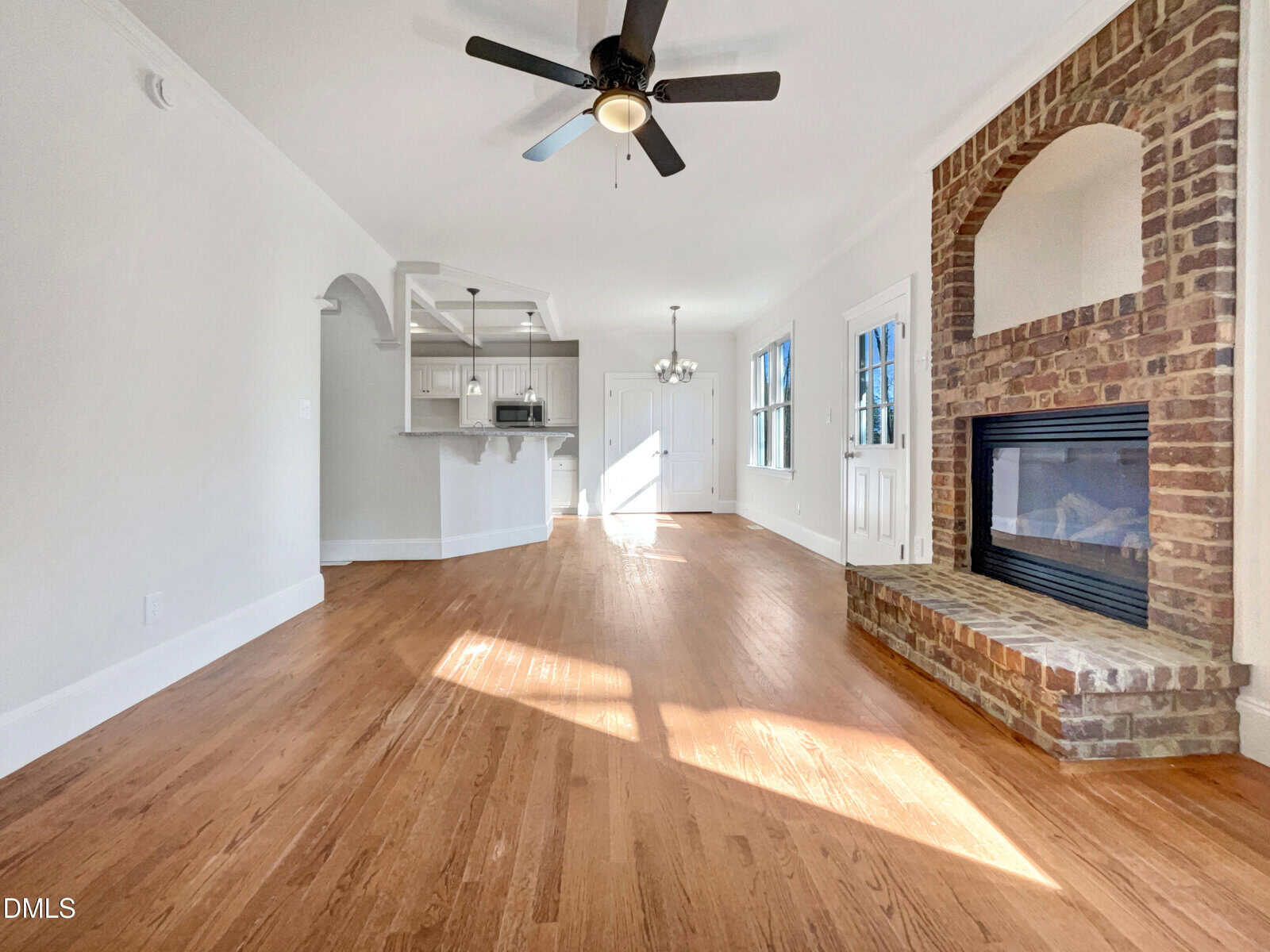 Bright living room with hardwood floors, brick fireplace, ceiling fan, and open view into a white kitchen. Large windows and glass door let in natural light, creating a warm, inviting space.