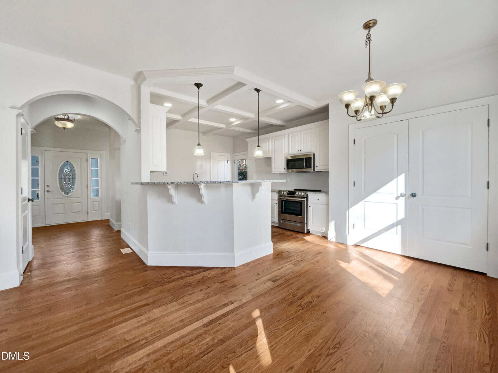 Bright, open kitchen and dining area with hardwood floors, white cabinets, stainless steel appliances, breakfast bar with pendant lights, double doors, and arched entryway leading to a front door with decorative glass.