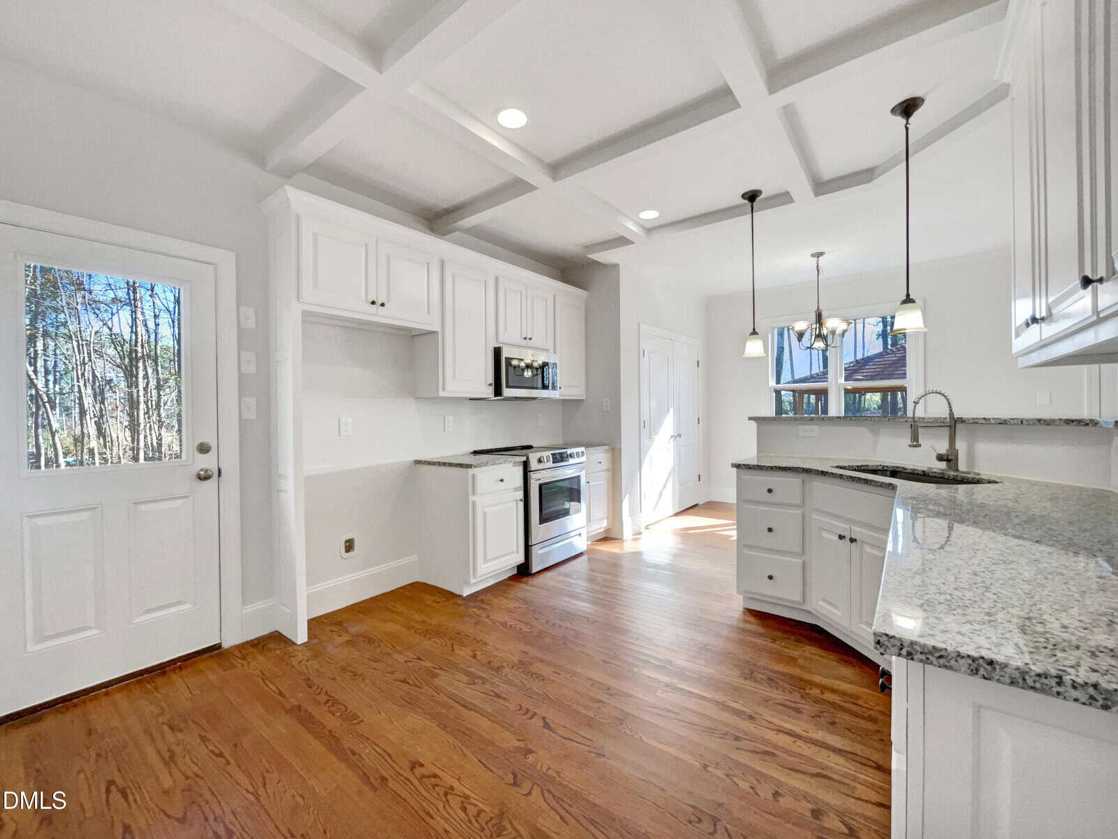 Bright kitchen with white cabinets, granite countertops, stainless steel appliances, and wood flooring. A door and large window let in natural light; pendant lights hang over a curved island with a sink.