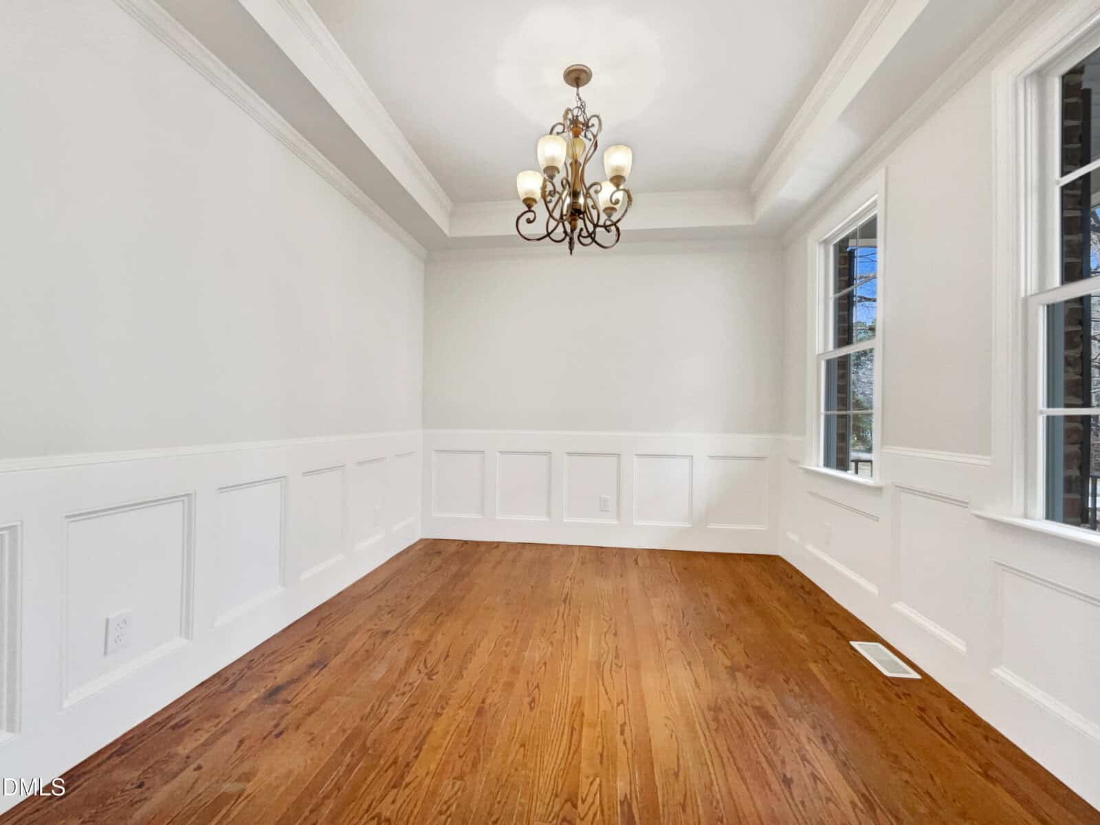 A bright, empty dining room with white wainscoting, hardwood floors, a tray ceiling, and an ornate chandelier. Large windows on the right wall let in natural light.