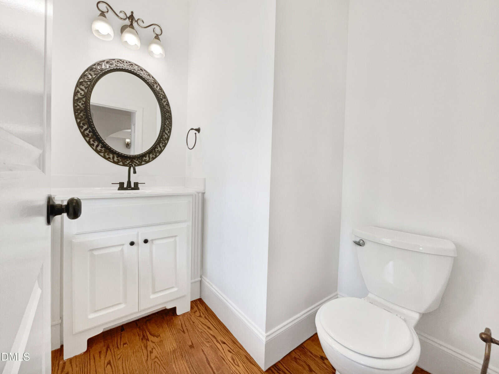 A small bathroom with a white vanity and ornate round mirror, a light fixture with three bulbs above, white walls, wood floor, and a white toilet to the right.