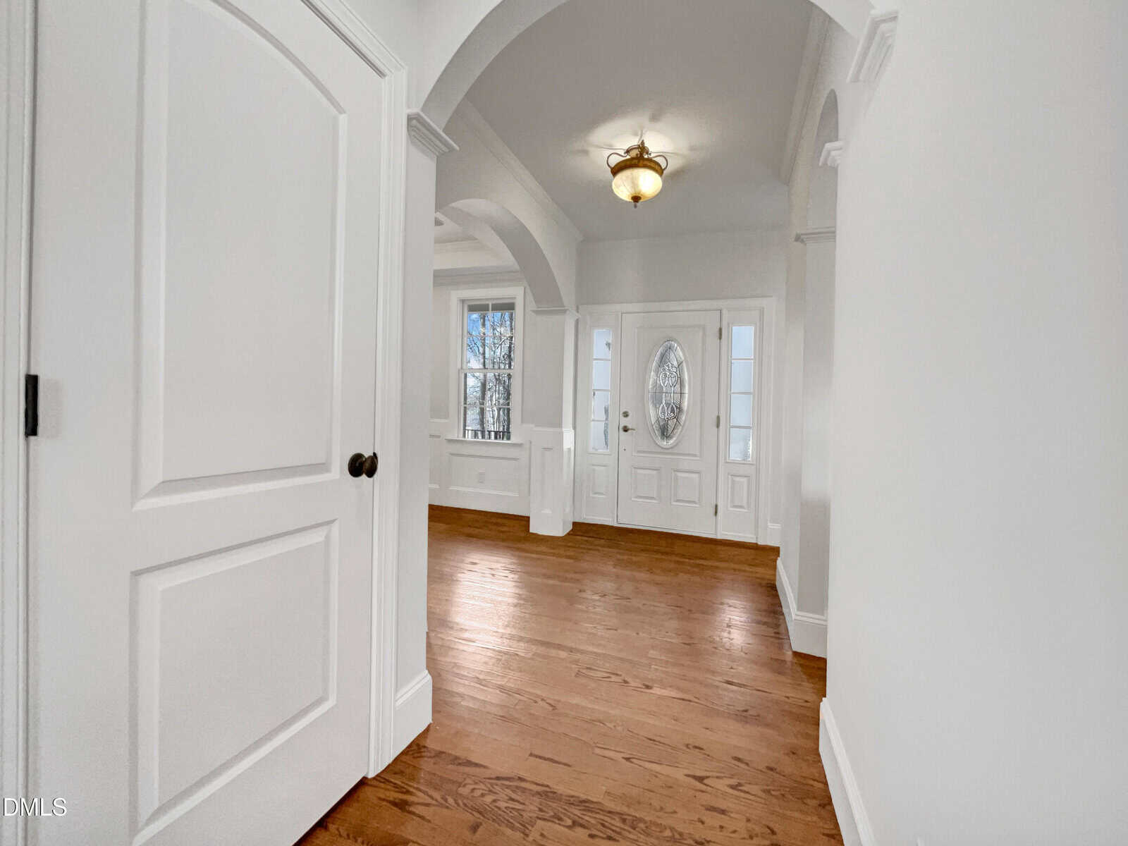 Bright entryway with hardwood floors, white walls, a round light fixture, arched doorways, and a front door with an oval glass window flanked by two narrow windows.