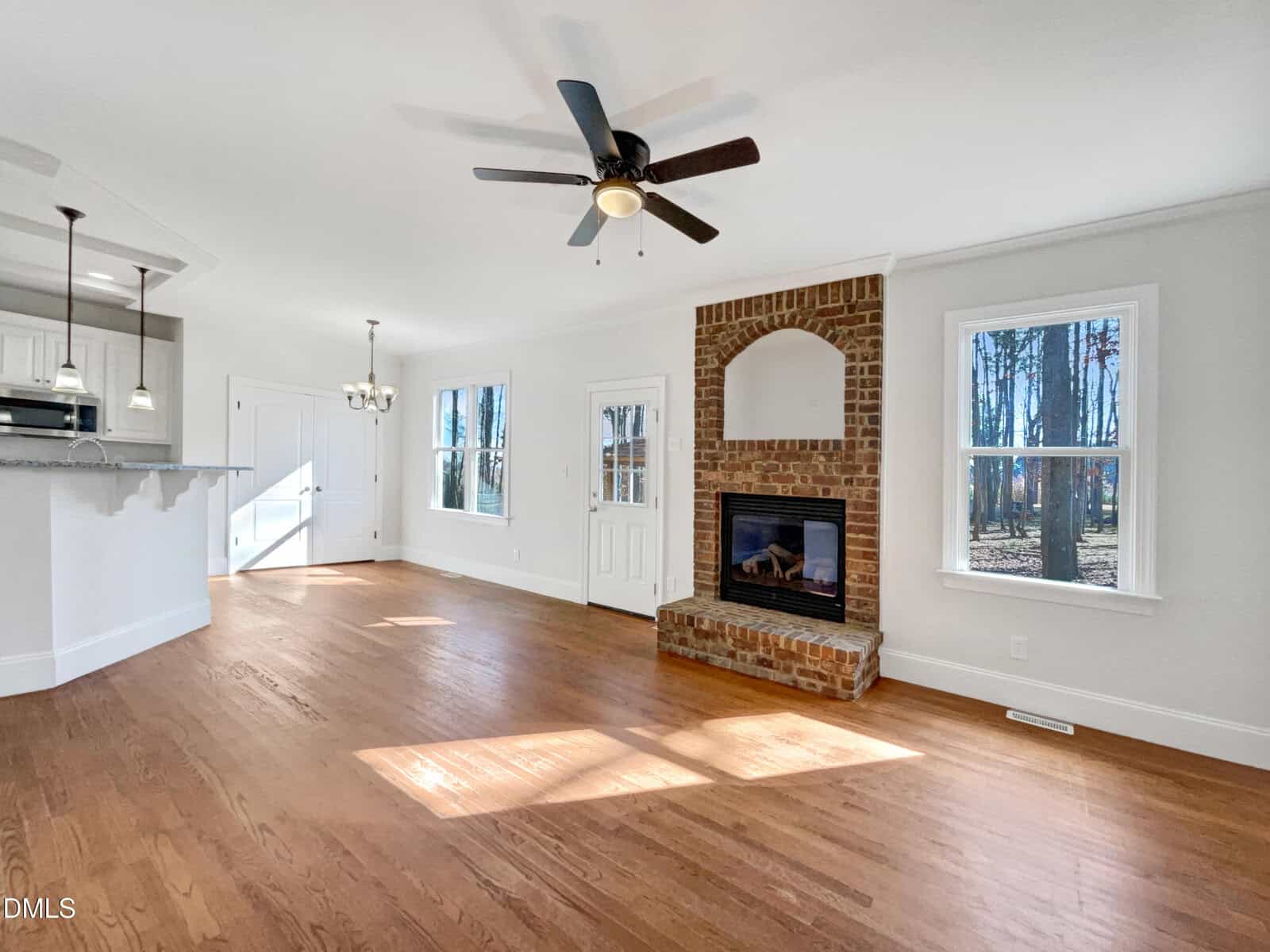 Bright living area with hardwood floors, a brick fireplace, white walls, large windows, ceiling fan, and open view to a white kitchen with bar counter and hanging lights. Sunlight streams in through the windows.