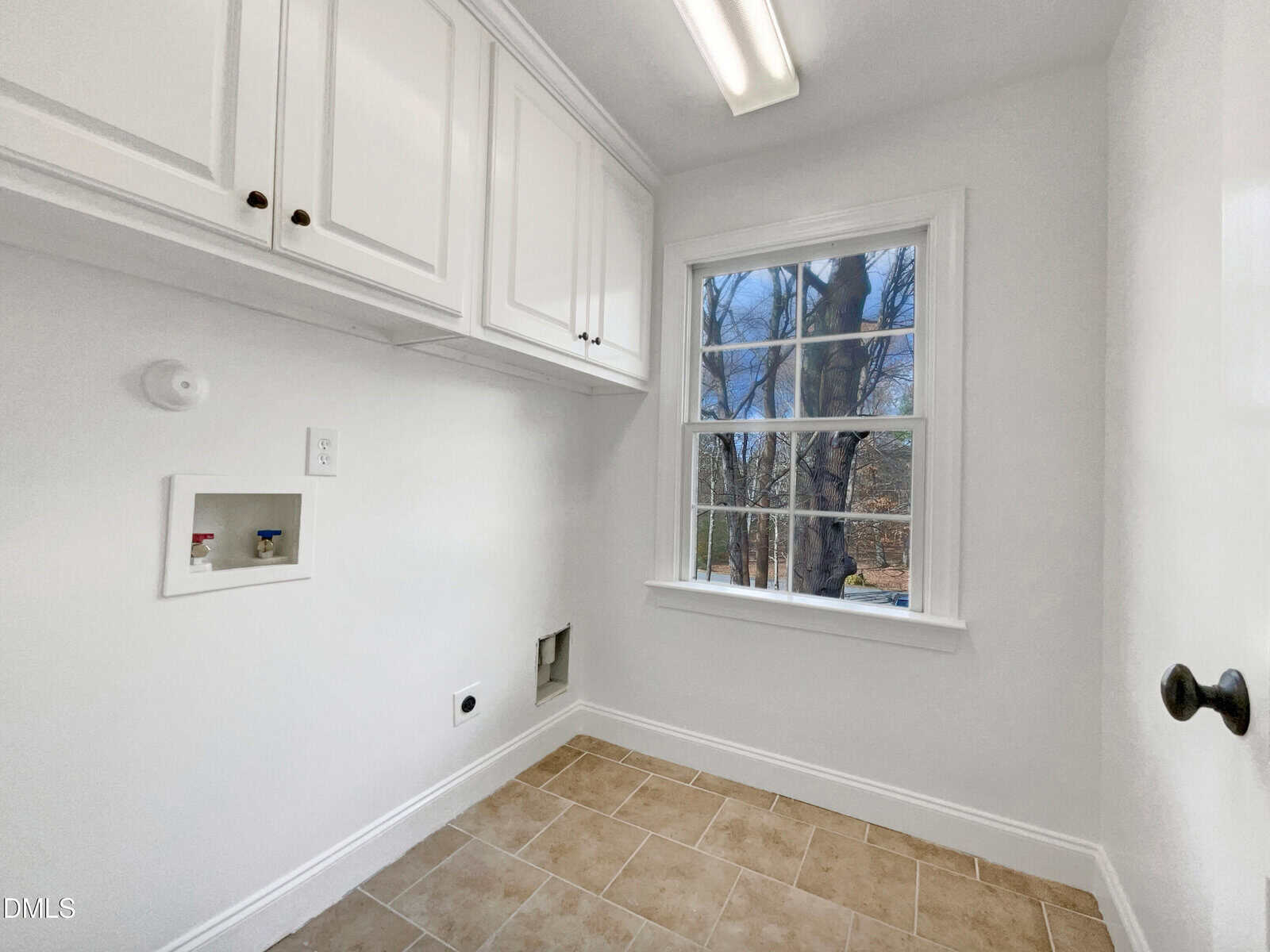 A bright laundry room with white cabinets, a window showing trees outside, tan tile floor, and hook-ups for a washer and dryer.