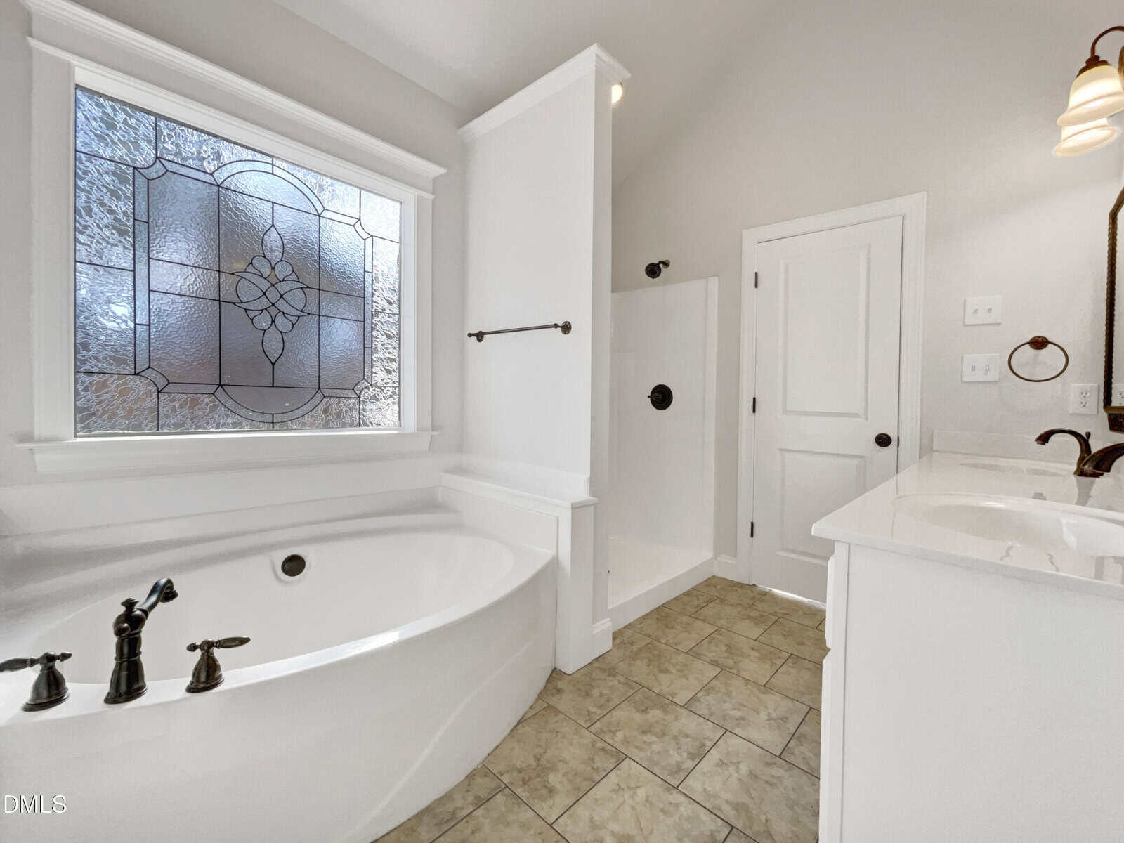 A bright bathroom with a large oval bathtub, a walk-in shower, double sink vanity, and a decorative frosted window. The space features neutral tile flooring and white walls.
