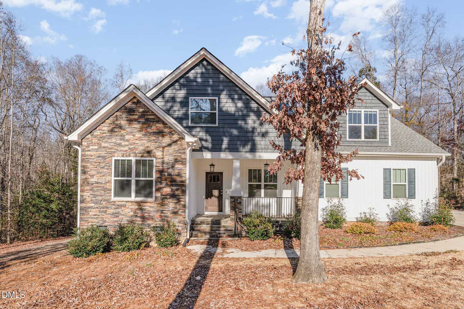 A two-story suburban house with a stone and dark siding exterior, white trim, covered porch, and landscaped front yard sits elegantly at 100 Lexington Avenue in Zebulon. A leafless tree stands in front, and the background shows tall, bare trees under a partly cloudy sky.