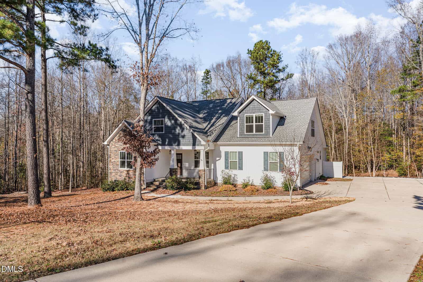 A two-story house with gray roof, white siding, and green shutters sits near a wooded area. There is a long concrete driveway, leaf-covered ground, and bare trees surrounding the property under a partly cloudy sky.