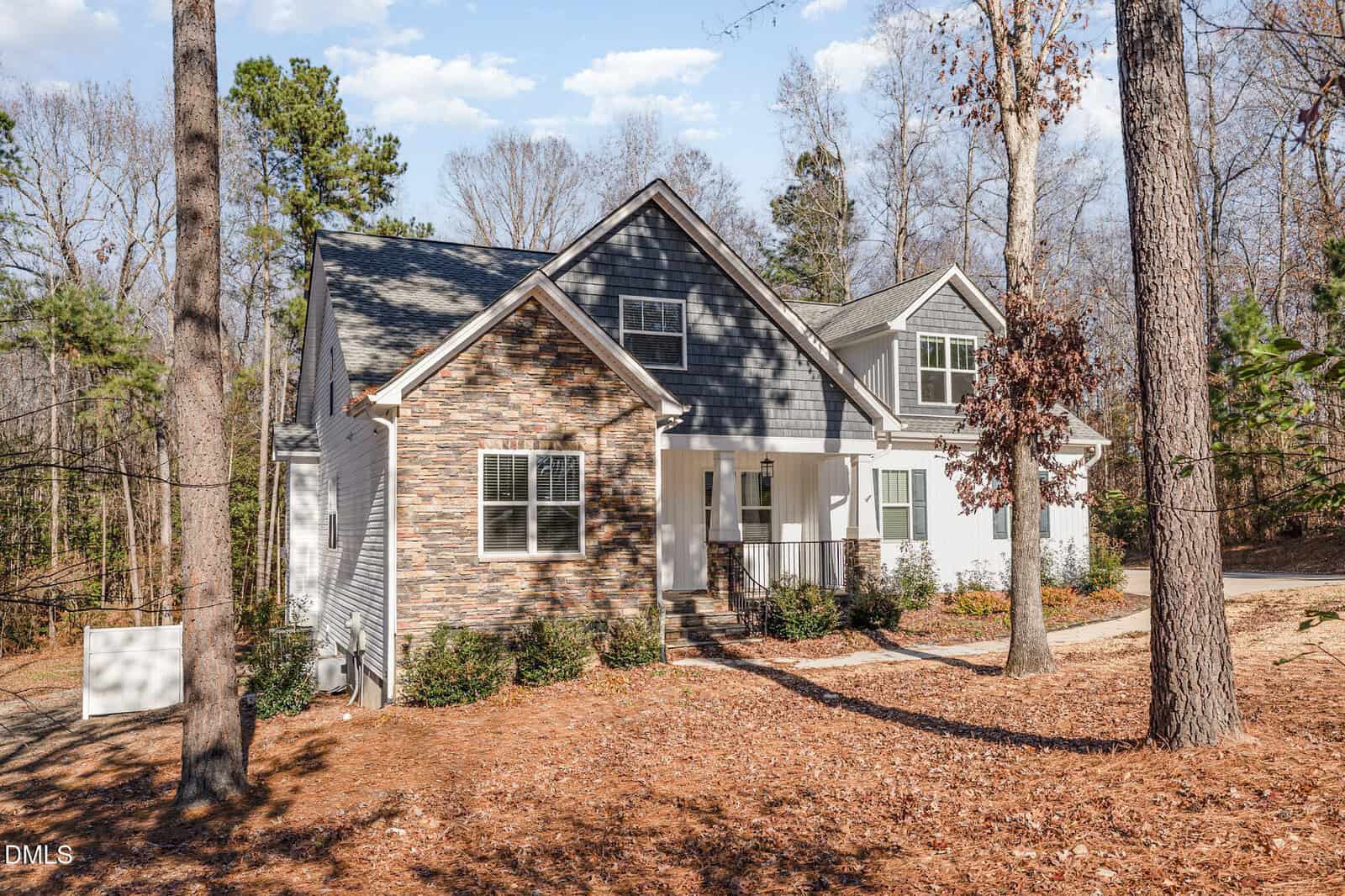 A modern house with stone and white siding, black shutters, and a covered porch sits among tall trees on a wooded lot with pine straw ground cover. The sky is partly cloudy.