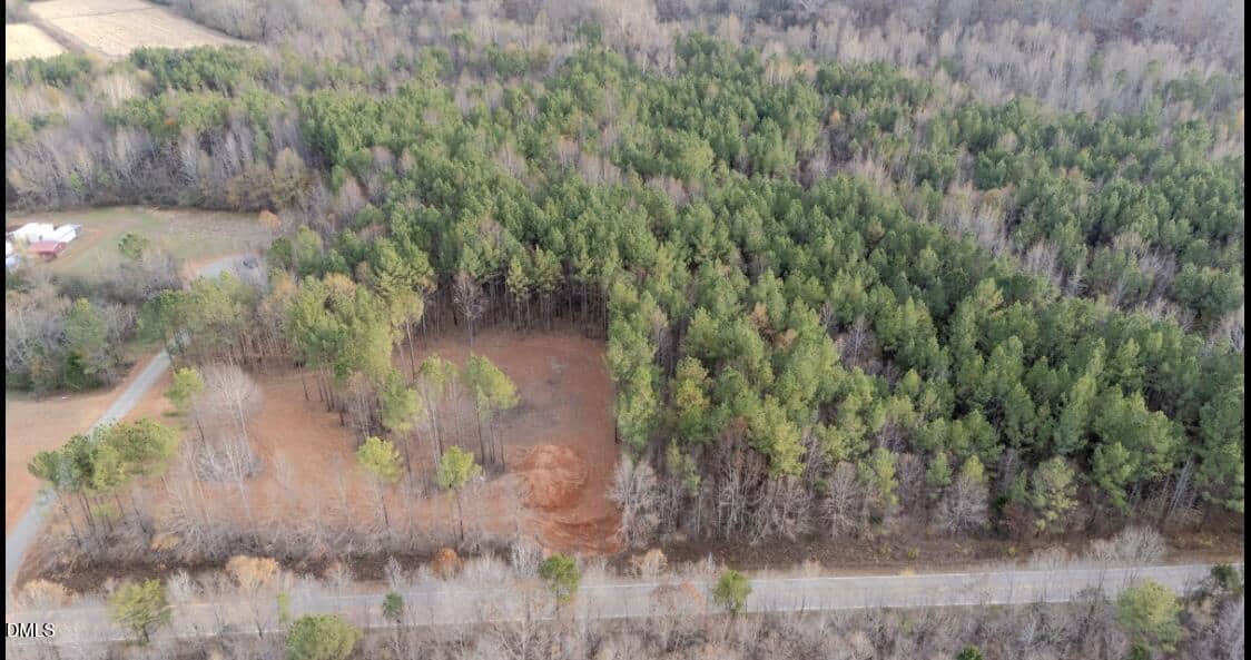 Aerial view of a forested area with a cleared patch of reddish-brown soil in the center, surrounded by dense trees. A paved road runs along the bottom edge, and an open field is visible in the upper left corner.