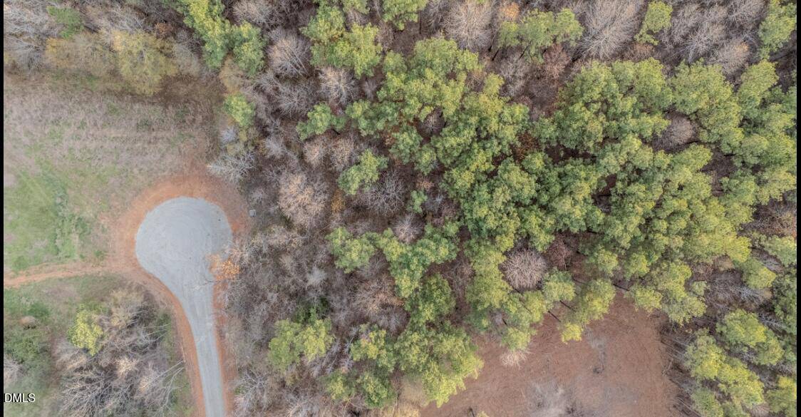 Aerial view of a cul-de-sac at the edge of a forested area, with green trees and brown foliage surrounding a circular paved road. The left side shows a grassy open space.