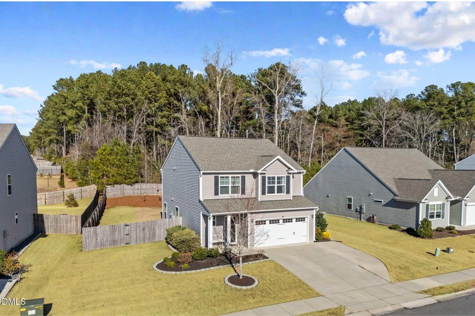 A two-story gray house with white trim and an attached garage sits on a well-kept lawn, surrounded by a wooden fence and neighboring homes, with trees and a blue sky in the background.