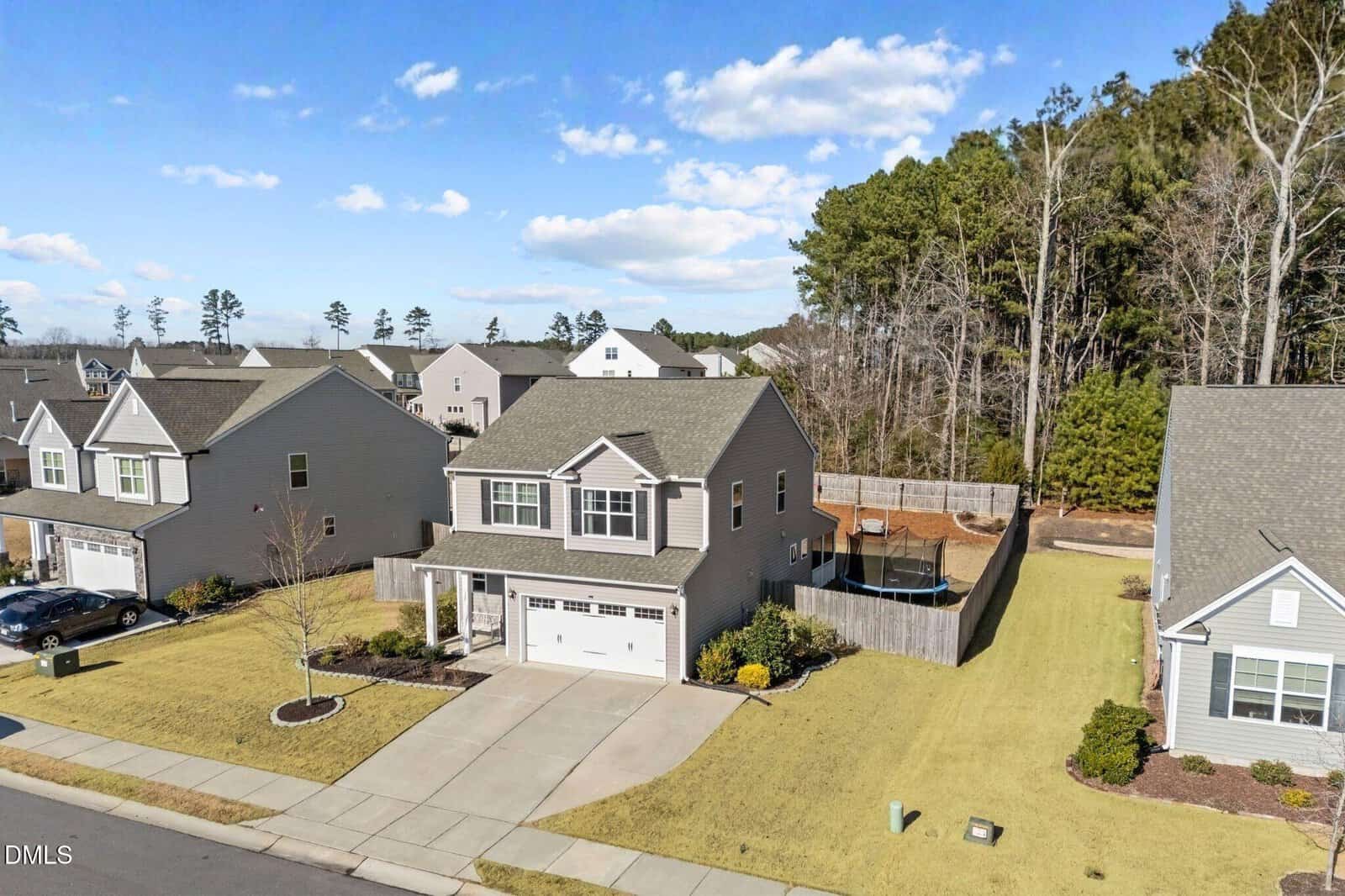 A two-story suburban house with a front porch, attached garage, fenced backyard, and trampoline; neighboring houses and trees line the street under a blue sky with scattered clouds.
