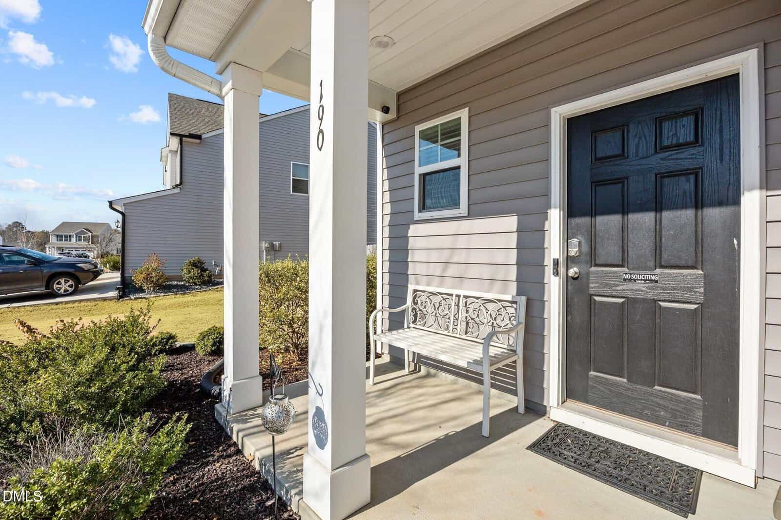 A front porch with a black door, a white decorative bench, white pillars, gray siding, and landscaping with bushes. A neighboring house and a parked car are visible in the background under a blue sky.