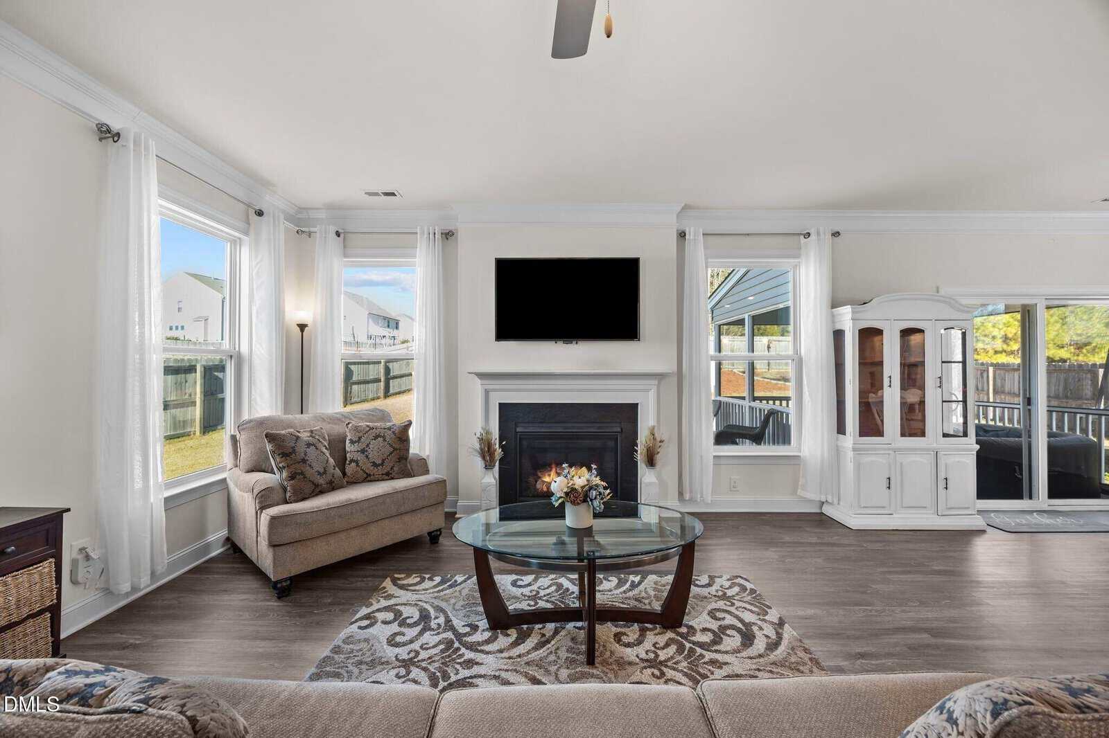 Bright living room with large windows, a glass coffee table, patterned rug, cushioned chair, white fireplace with a mounted TV above, and a white display cabinet. Neutral tones and natural light fill the space.