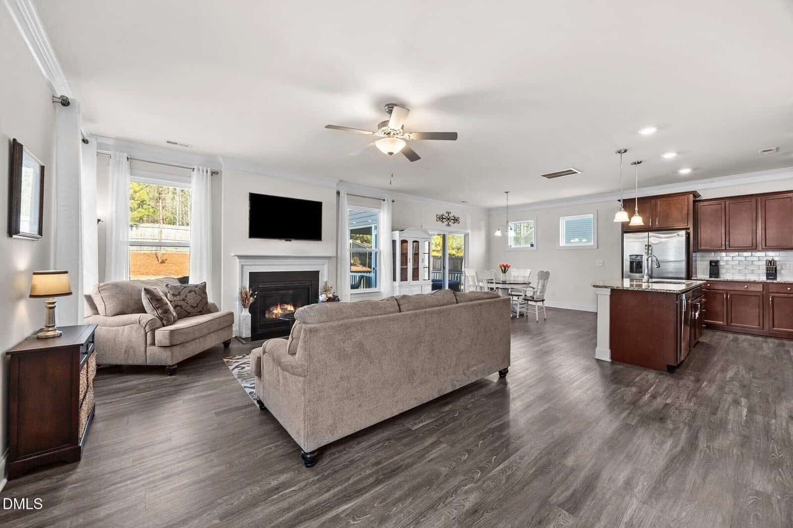 Open-concept living room and kitchen with dark wood floors, beige sofas, fireplace, wall-mounted TV, stainless steel appliances, and large windows letting in natural light.