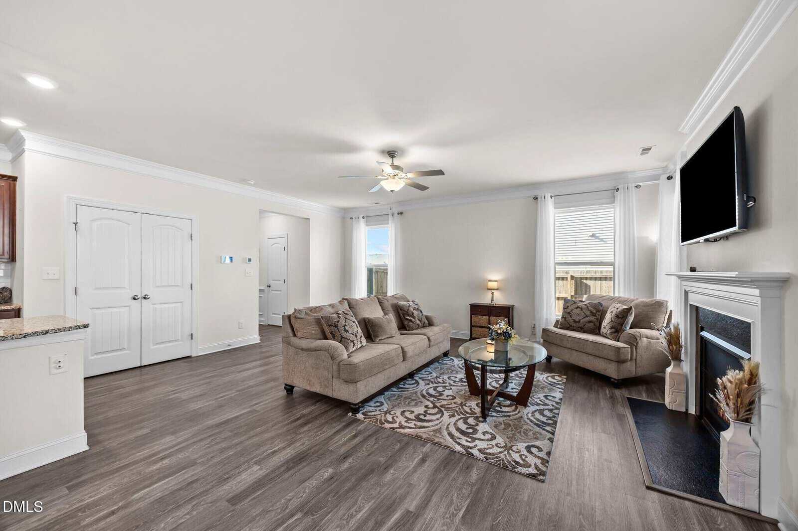 Bright living room with two beige sofas, a glass coffee table, rug, fireplace with TV above, side table, wood floors, and large windows with white curtains letting in natural light.