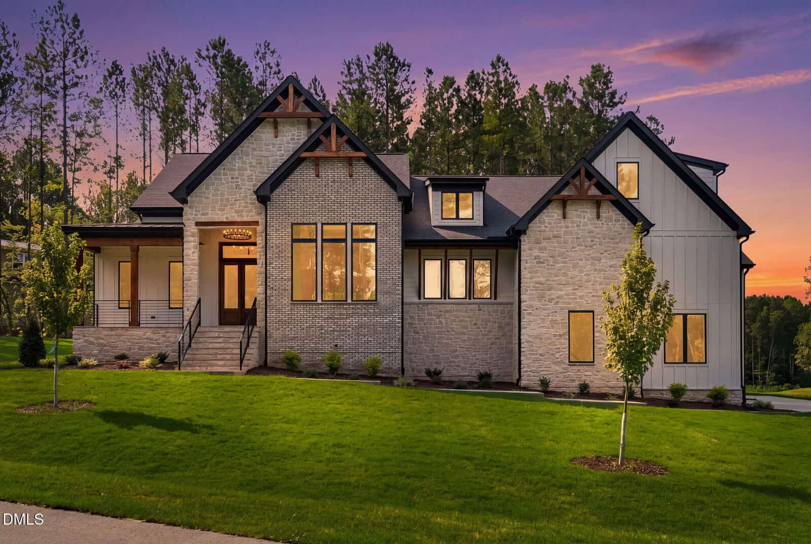 Exterior of a modern craftsman home at dusk with stone and brick facade, wood accents, black-trimmed windows, covered porch, lush green lawn, and pine trees under a purple-orange sunset sky.