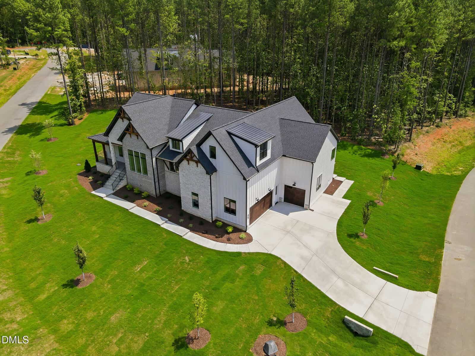 A large modern house with gray siding, black roof, and wood trim sits on a spacious green lawn, surrounded by young trees and bordered by a curved driveway and dense forest.