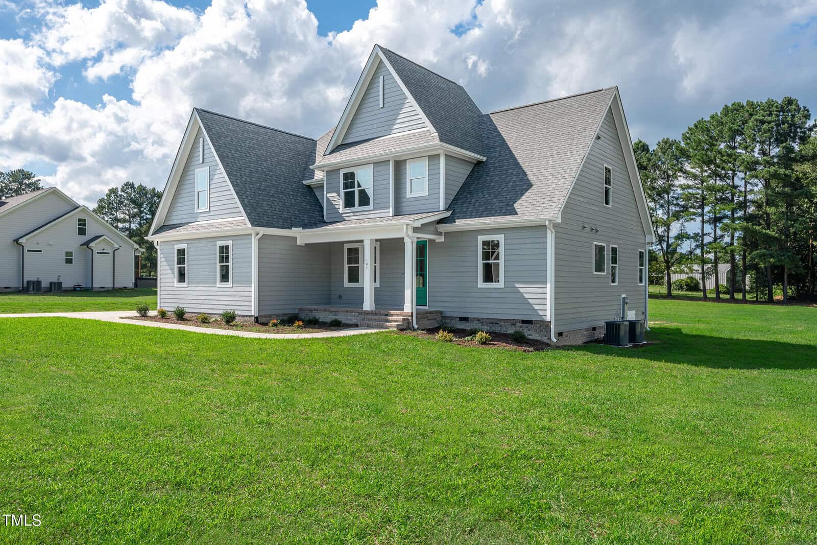 A two-story gray house with white trim and a covered front porch stands on a large, well-kept lawn. The house has multiple gables, an attached garage, and is surrounded by trees and a clear sky with clouds.