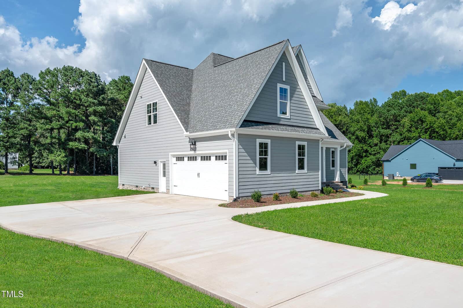 A modern gray house with white trim and a two-car garage sits on a large, well-maintained lawn. The driveway is wide and concrete, and there are trees and another house in the background under a partly cloudy sky.