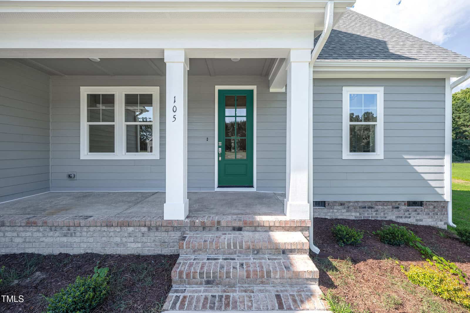 The image shows the front entrance of a modern house with light gray siding, a green door with glass panels, white trim, brick steps, and a small covered porch. The house number 105 is visible on a column.