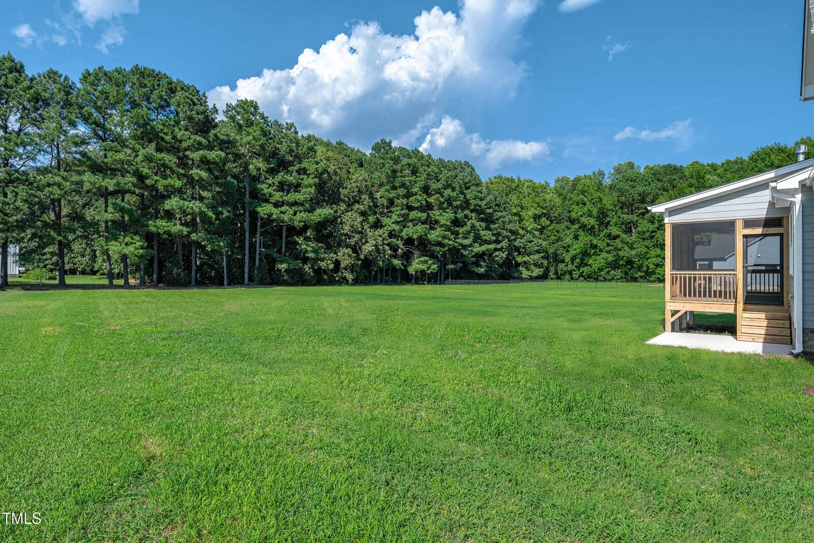 A large, grassy backyard with a line of dense trees in the background; part of a house with a small screened porch is visible on the right under a clear blue sky with some clouds.