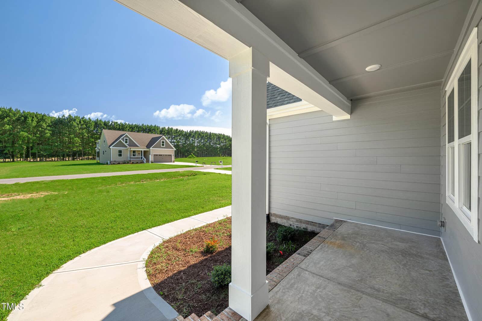View from a covered front porch with gray siding, looking out onto a green lawn, a curved walkway, and another house in the distance with surrounding trees.