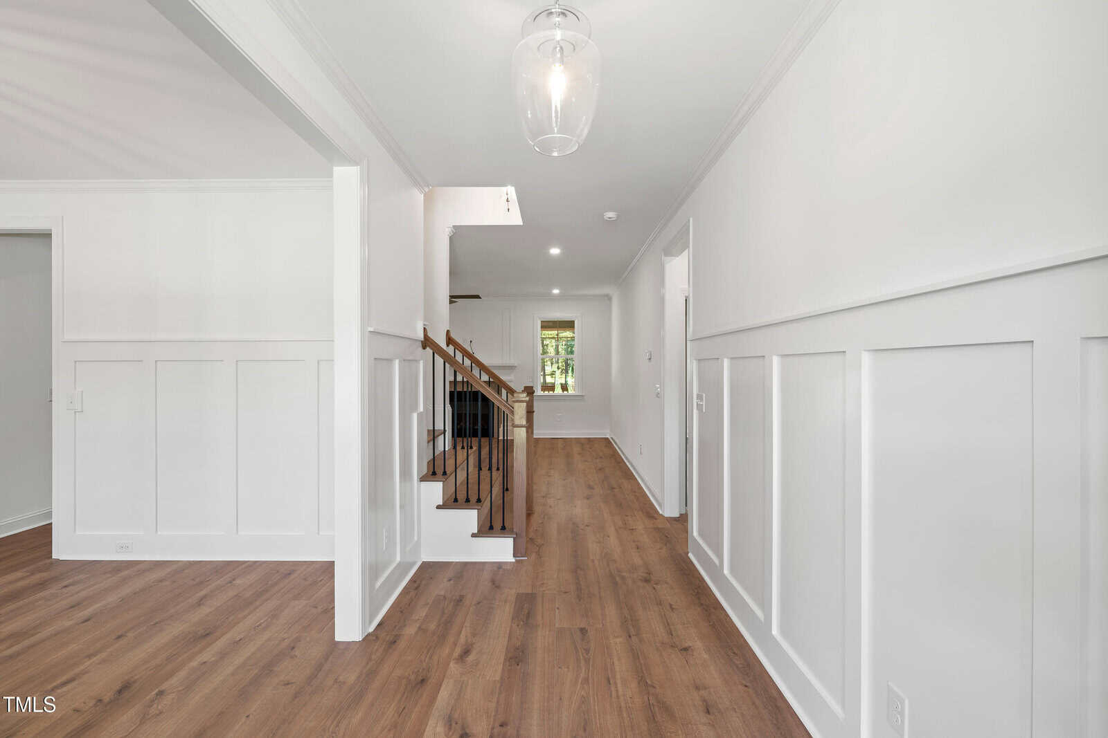 Bright entryway with white walls, wainscoting, wood floors, and a staircase with a wooden handrail. A glass light fixture hangs from the ceiling. Natural light comes from a window at the far end.