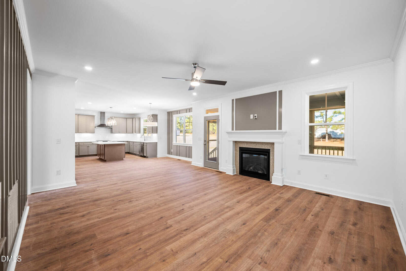 Spacious open-concept living area with wood flooring, ceiling fan, fireplace, and large windows. Kitchen with island and pendant lighting is visible in the background. Patio doors open to a fenced backyard.