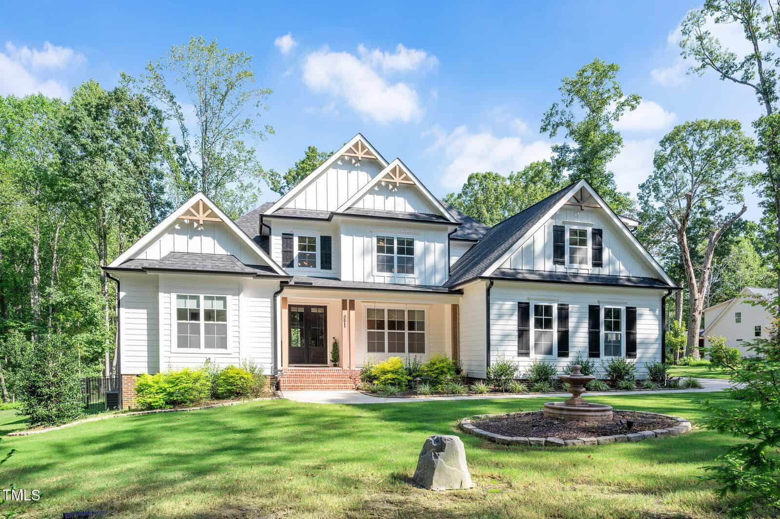 A large two-story white house with black trim and multiple gables sits on a green lawn at 3511 Bruce Garner Road, Wake Forest, surrounded by shrubs, a circular stone feature, and trees under a blue sky.