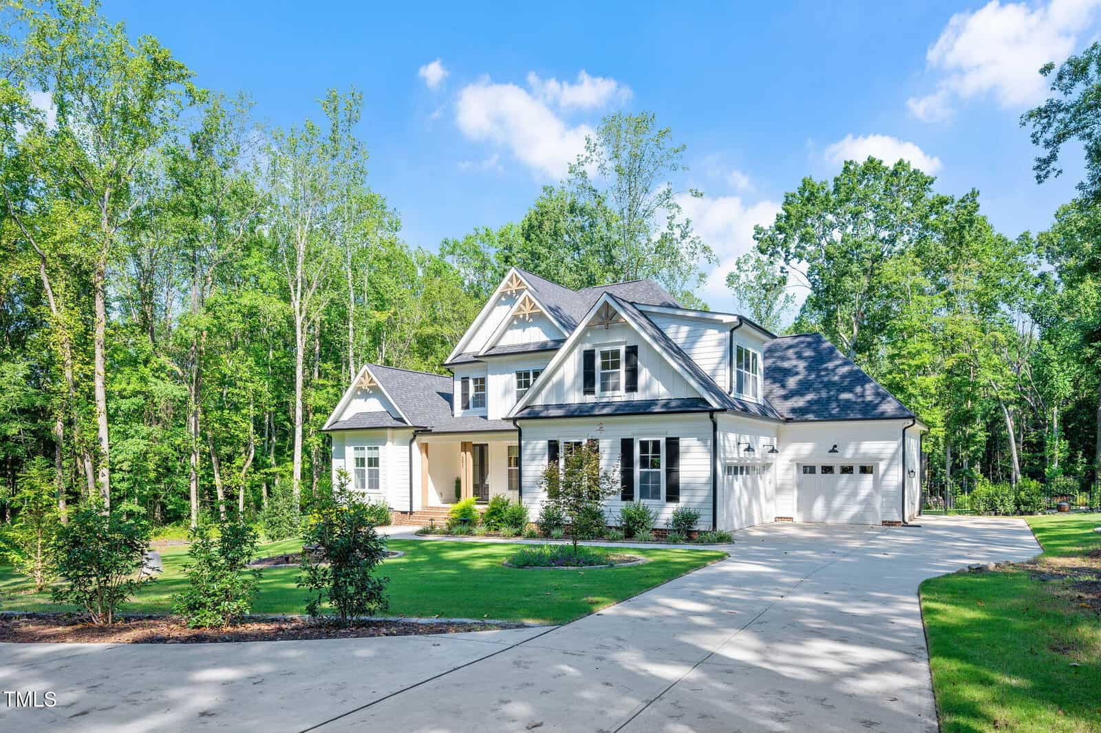 A two-story suburban house with white siding, black trim, and a covered front porch sits among tall trees. A wide driveway leads to a double garage, and the lawn is neatly landscaped under a blue sky with scattered clouds.