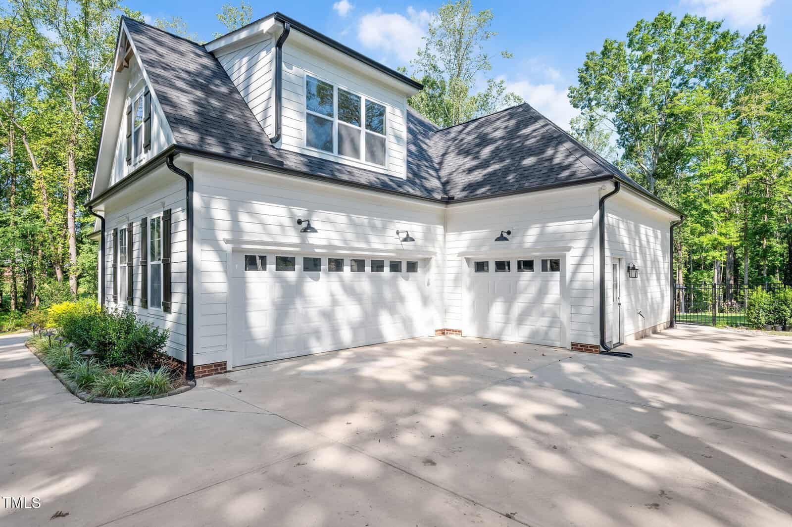 A white two-story house with black roof shingles features three garage doors and a concrete driveway, surrounded by trees and greenery under a blue sky.