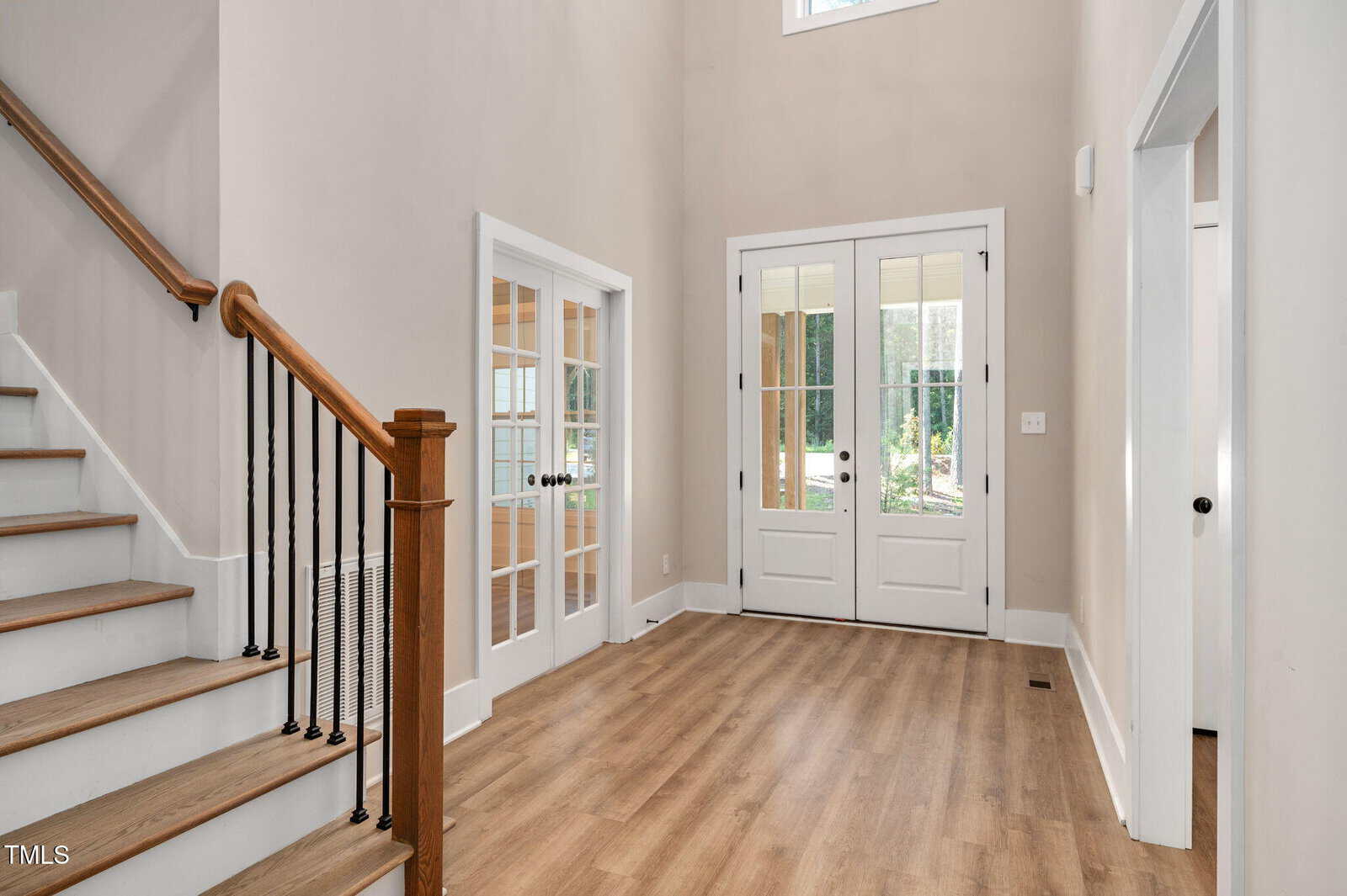 A spacious entryway with light wood flooring, double glass front doors, a staircase with a wooden handrail and black balusters, and a set of French doors leading to another room.