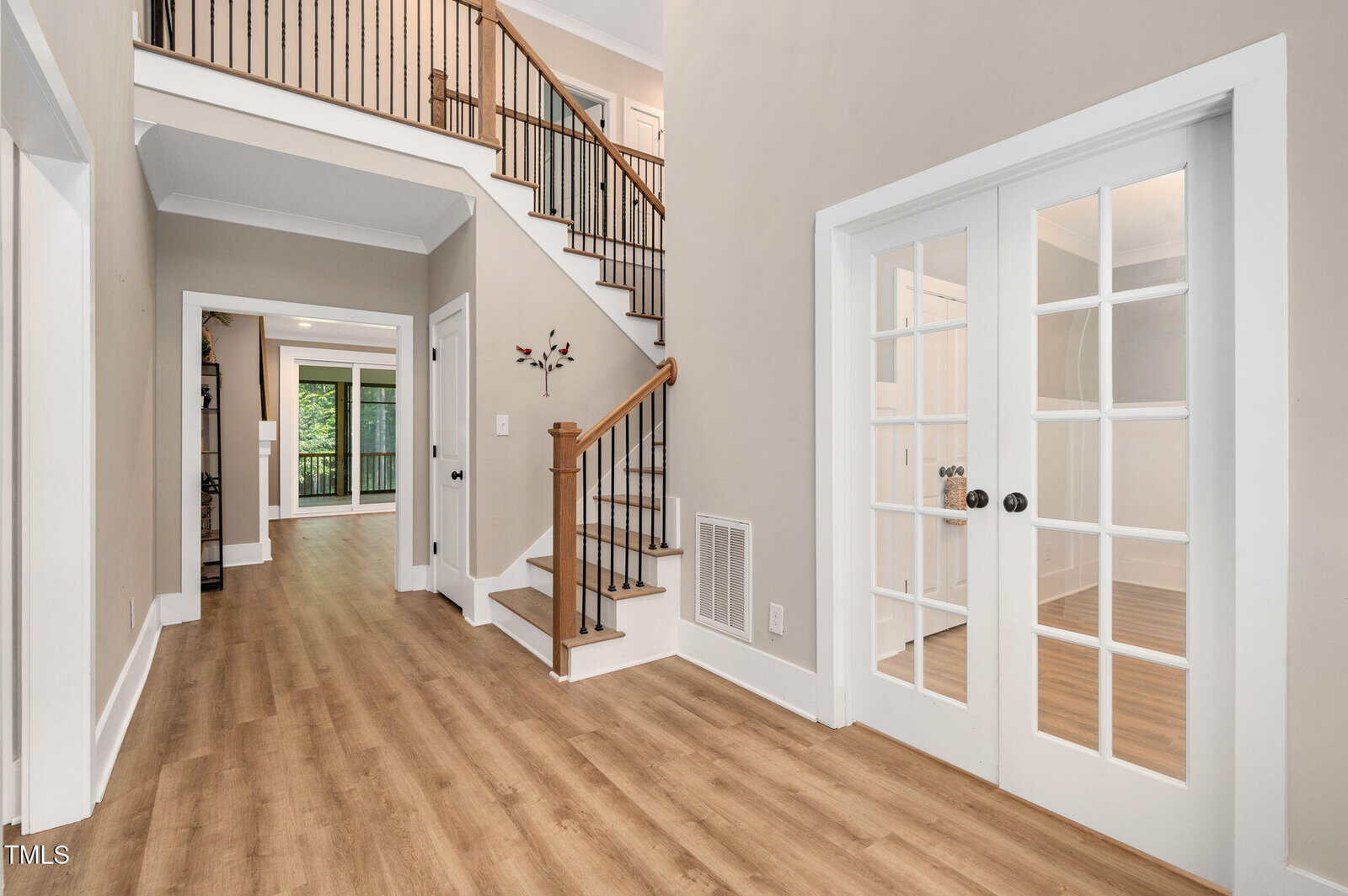 A hallway with light wood flooring, beige walls, white trim, a staircase with wooden handrails and black balusters, and double glass doors leading to a separate room. An open doorway reveals another room beyond.
