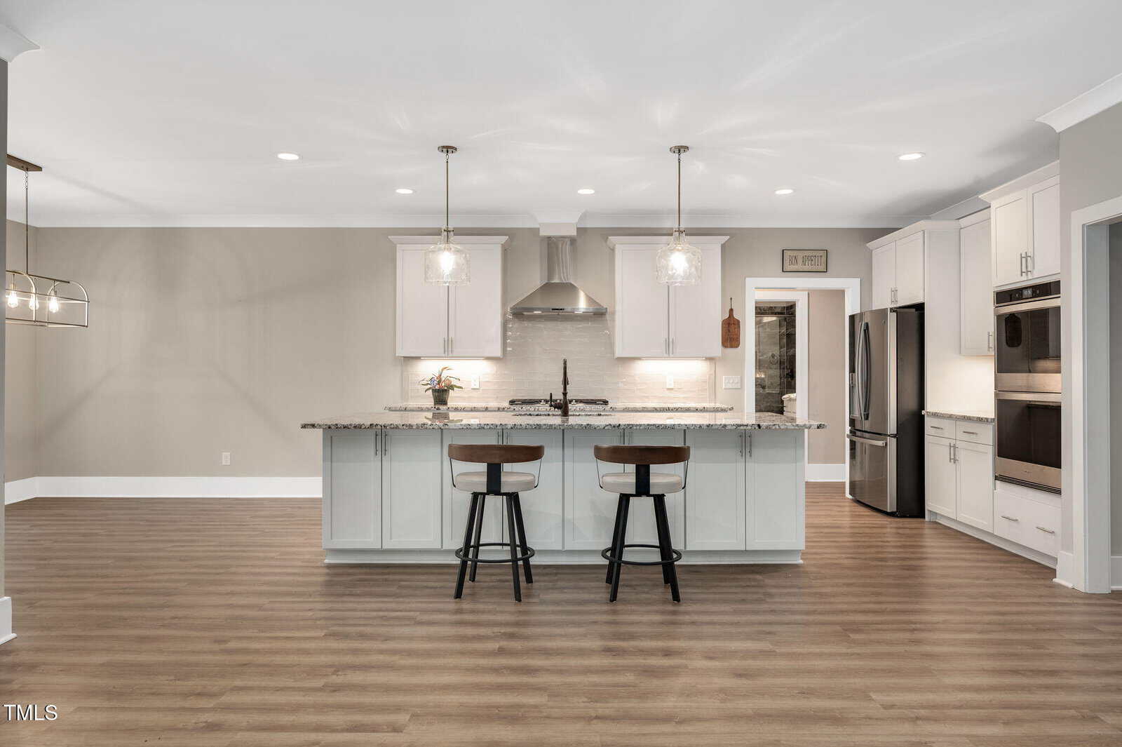A modern kitchen with white cabinets, stainless steel appliances, a large island with granite countertop, two barstools, pendant lights, and wood flooring. The space is open and well-lit.