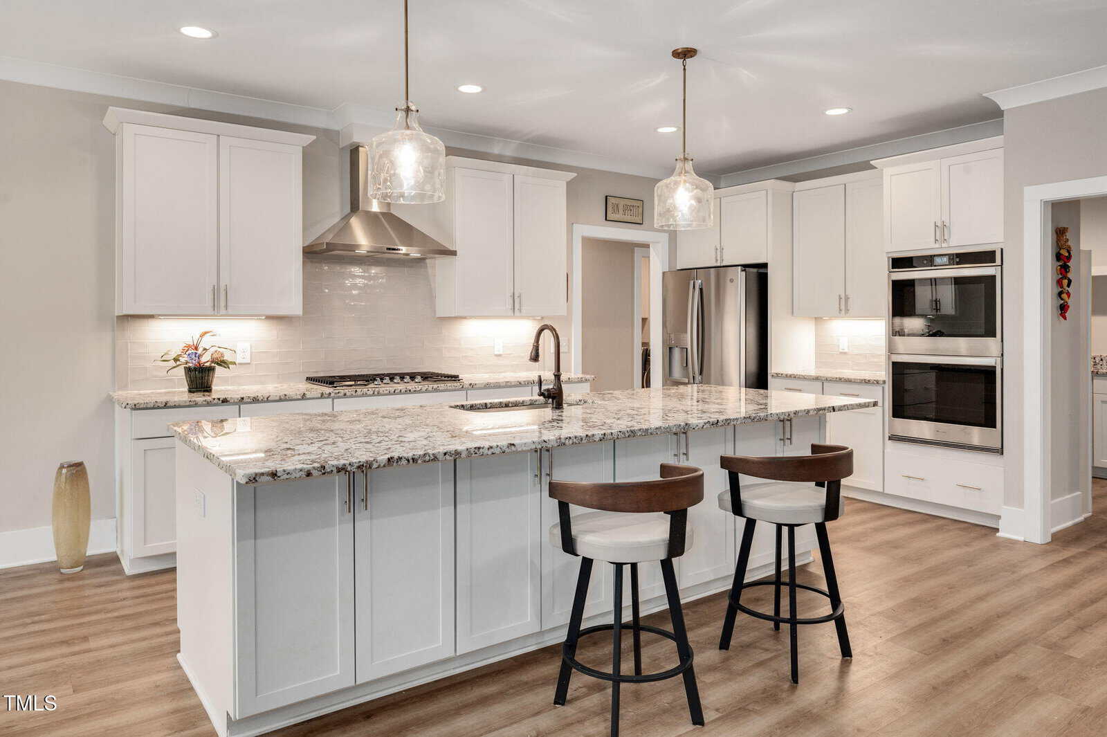 A modern kitchen with white cabinets, granite countertops, a center island with two chairs, stainless steel appliances including a double oven, and pendant lights hanging above the island.