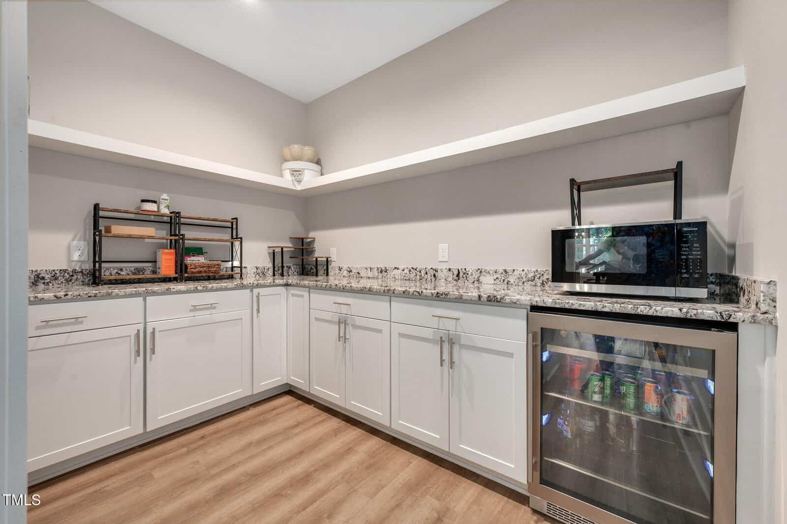 A kitchenette with white cabinets, granite countertops, light wood flooring, open shelves with decorative items, a microwave, and a glass-front mini fridge stocked with drinks.