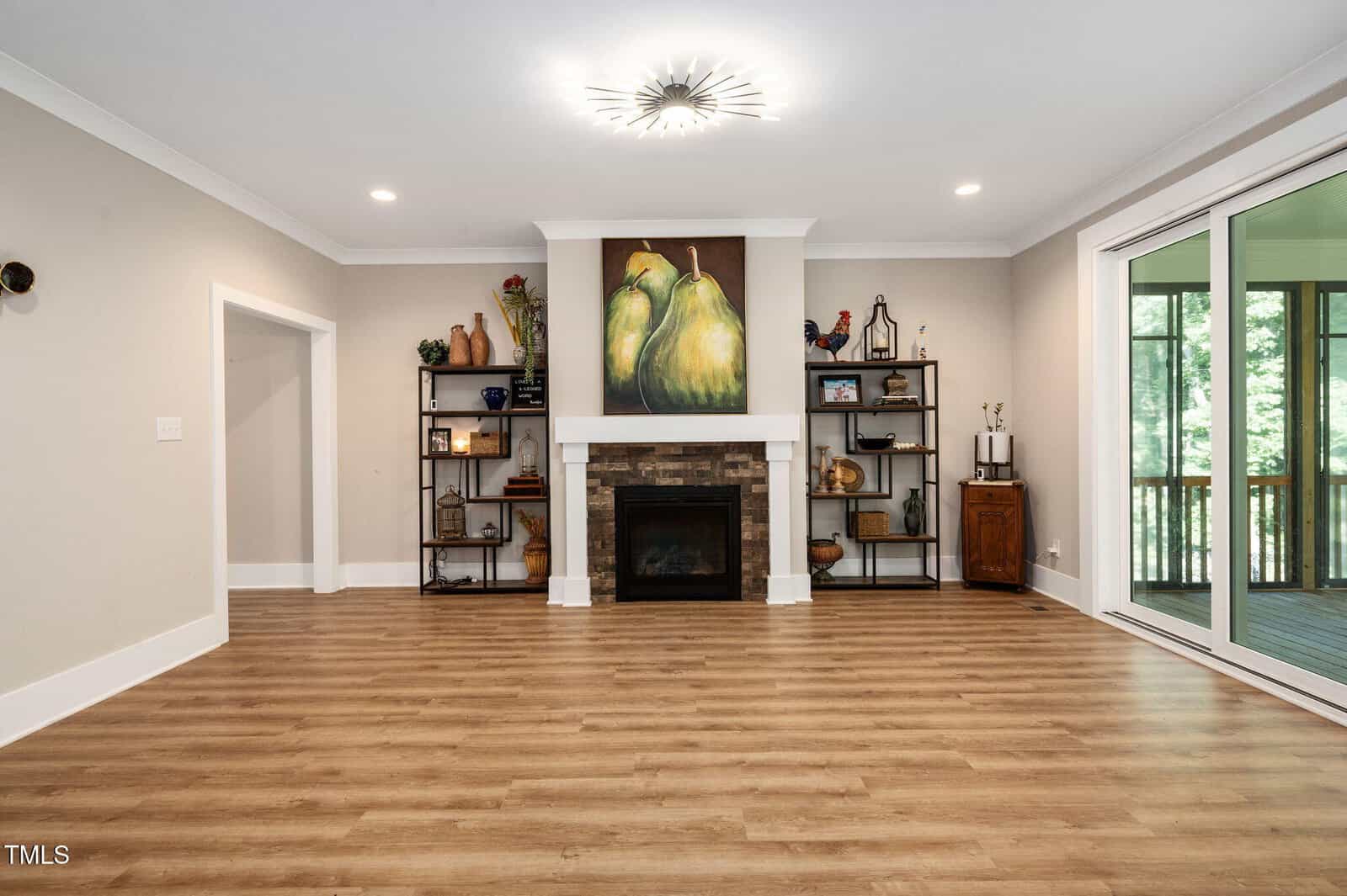 A spacious living room with light wood flooring, a fireplace centered on one wall, a painting of pears above the mantel, two shelving units with decorative items, and large glass doors opening to a screened porch.